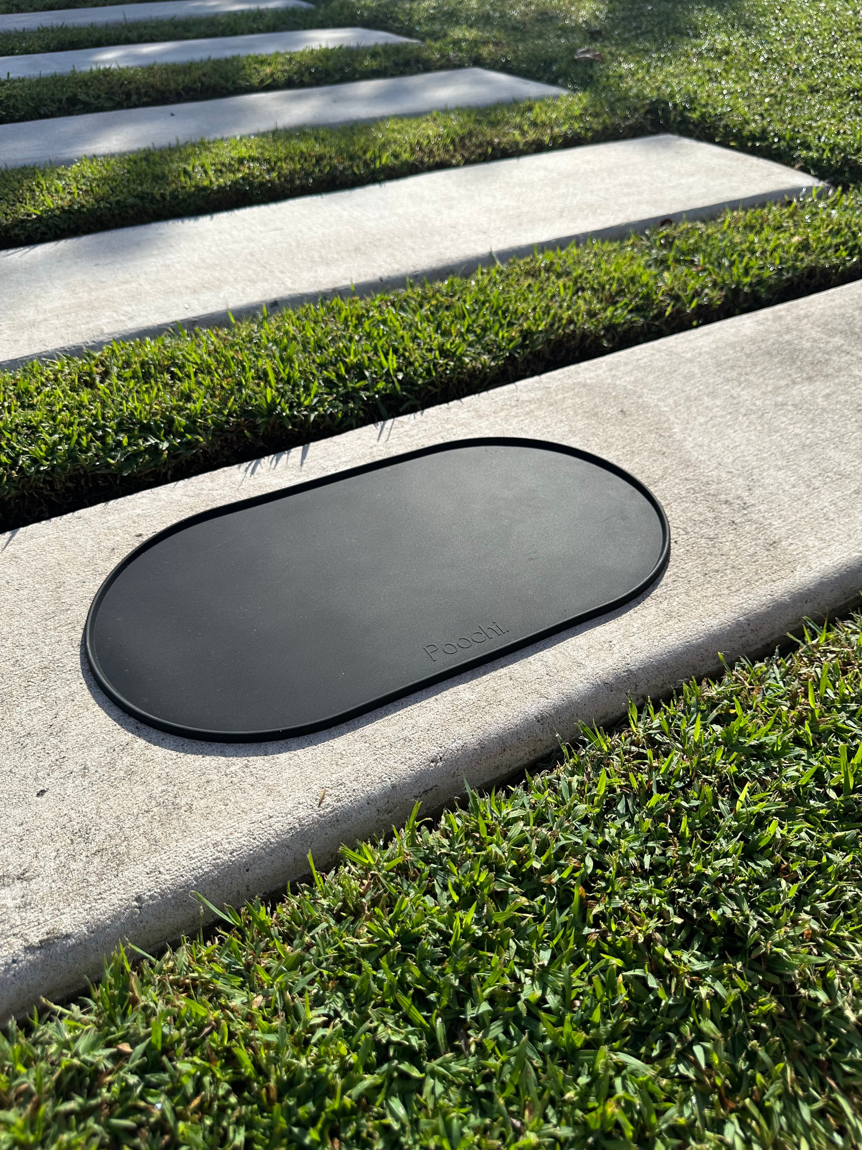 A black silicone feeding mat for pets such as dogs or cats, placed outside on the concrete in the sun, branded Poochi. 