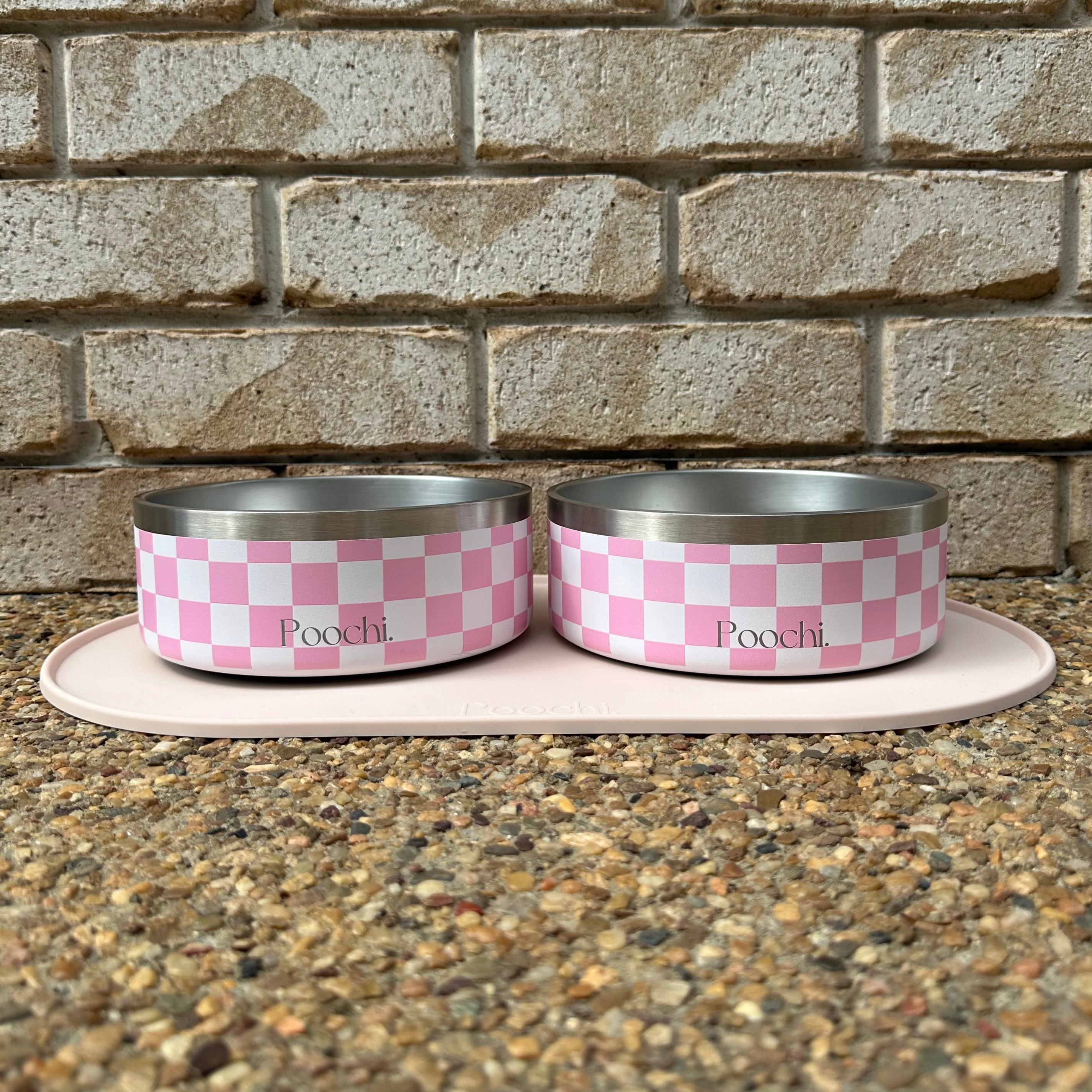 Two medium pink and white checkered stainless steel pet bowls with 'Poochi' branding on a stone surface against a brick wall.