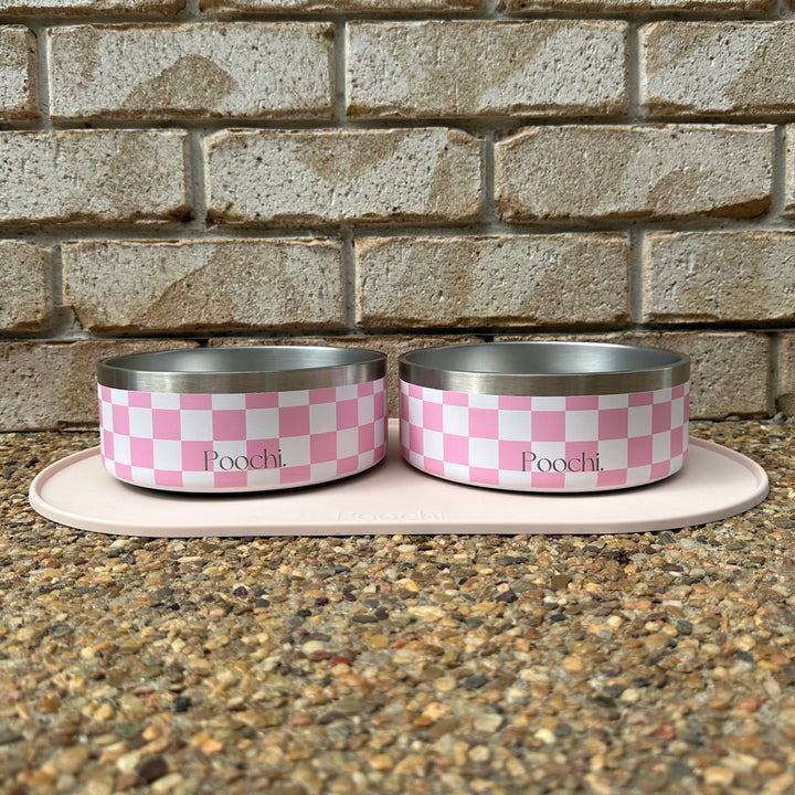Two medium pink and white checkered stainless steel pet bowls with 'Poochi' branding on a stone surface against a brick wall.