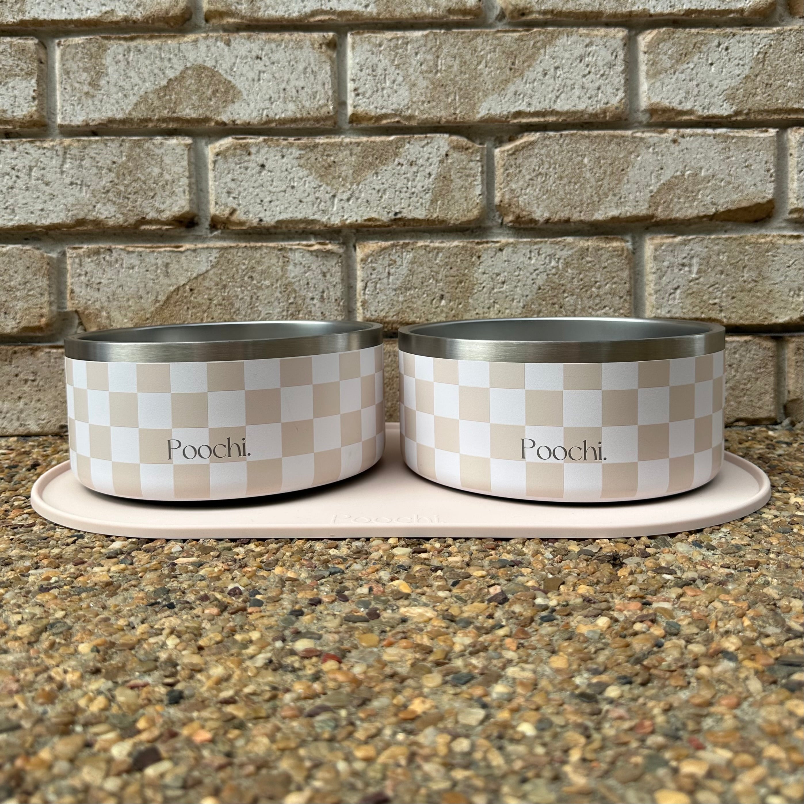 Two large checkered beige and white stainless steel pet bowls with 'Poochi' branding on a stone surface and brick wall background.