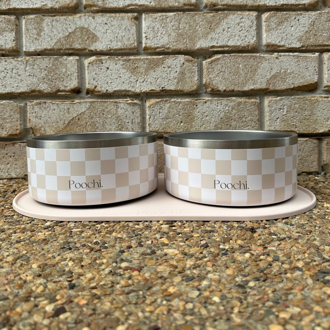 Two large checkered beige and white stainless steel pet bowls with 'Poochi' branding on a stone surface and brick wall background.