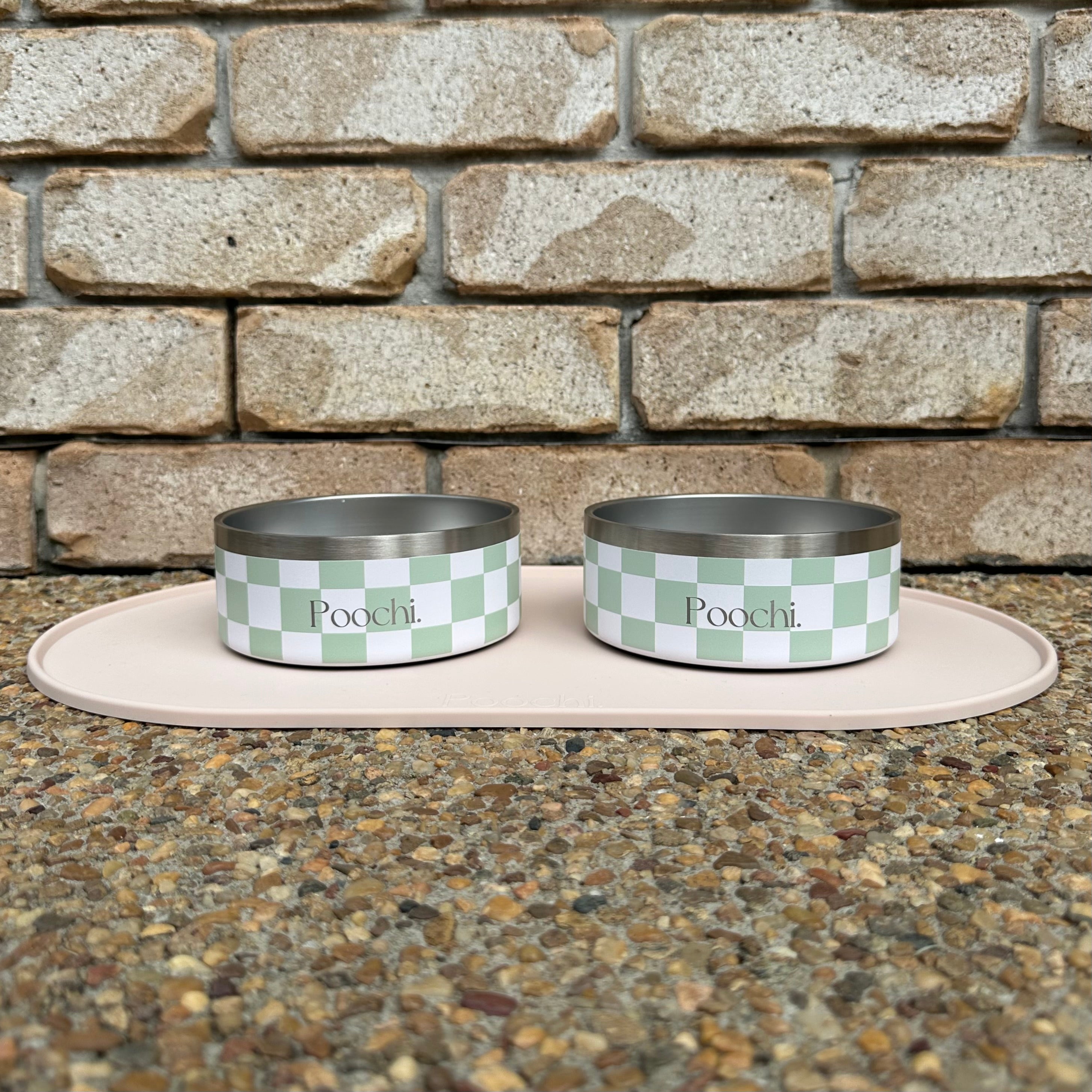 Two small green and white stainless steel checkered pet bowls with 'Pocchi' branding on a stone surface and brick wall background.